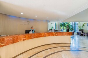 Ein Hotel-Empfangsbereich mit Marmor-Theke, einer Rezeptionistin und Blick ins Freie durch große Fenster.