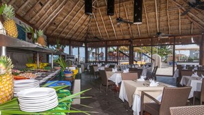 Un ristorante all'aperto con tetti di paglia, tavoli apparecchiati con tovaglie bianche e un'area buffet con frutta e bevande.