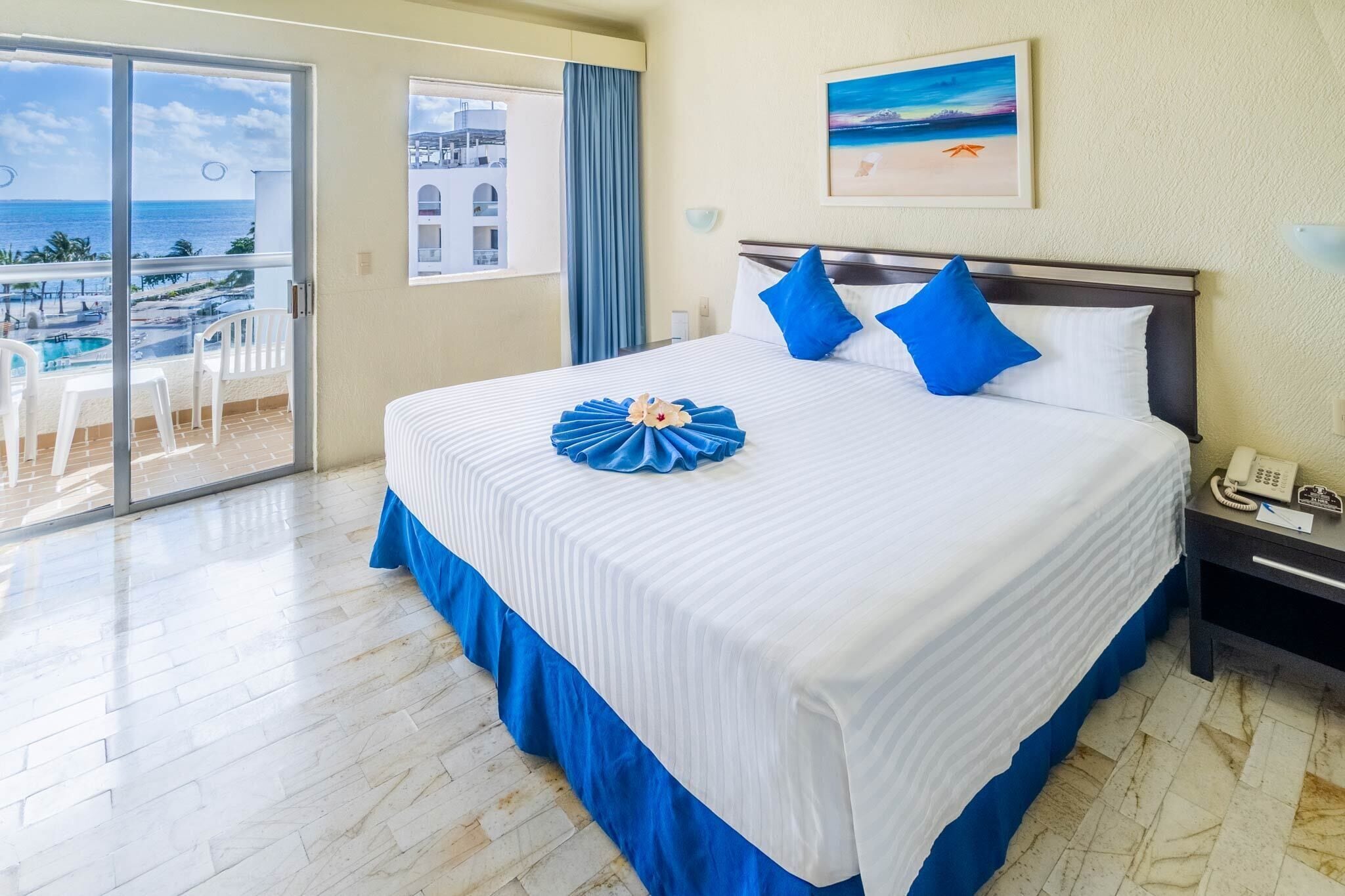 Habitación de hotel con una cama grande, vista al océano y un balcón con mobiliario exterior.
