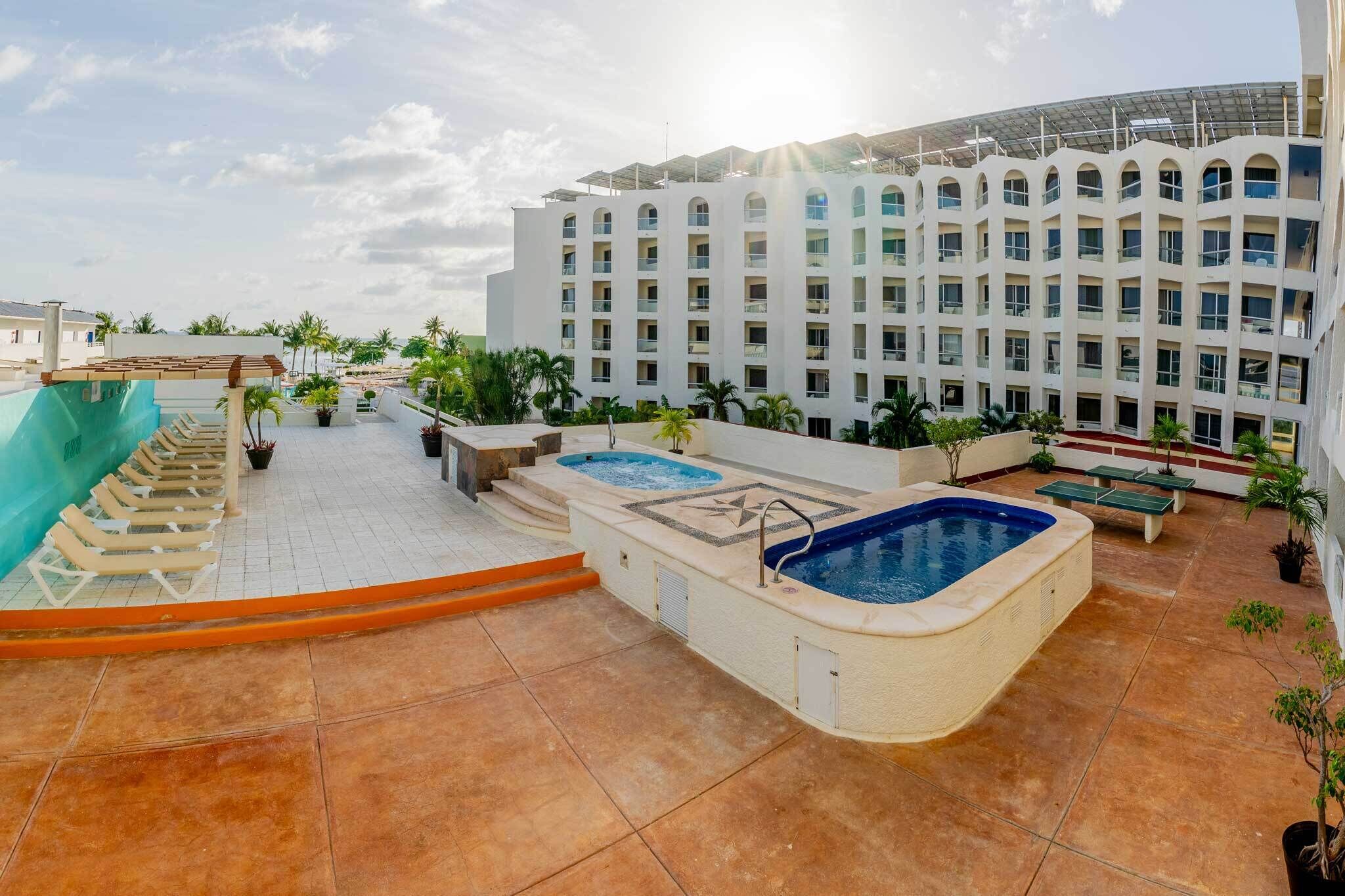 Un área de piscina en la azotea con varios jacuzzis y sillones de descanso, rodeada por un edificio de diseño moderno y un cielo despejado.