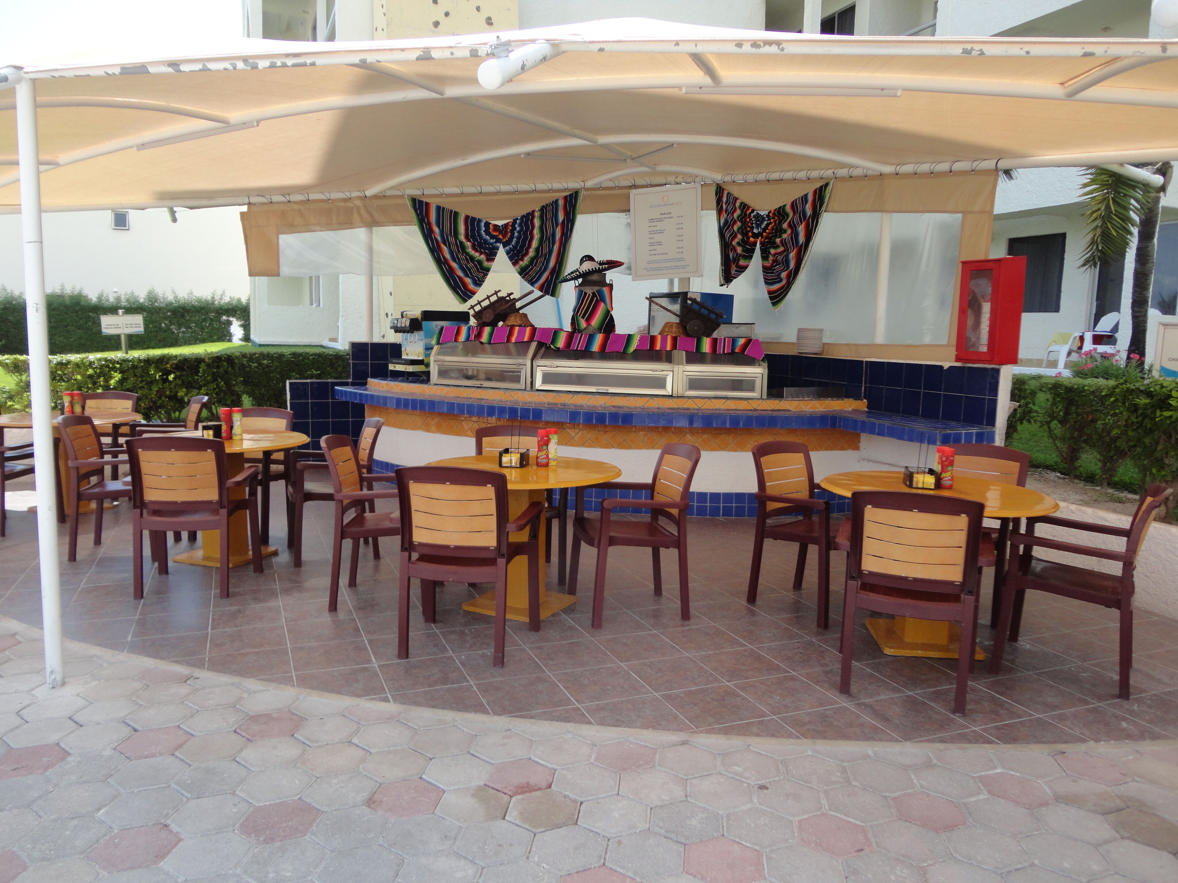 An outdoor cafe with hexagonal tiled flooring, wooden tables and chairs, a central counter with a menu, and a covered seating area.