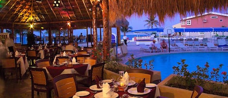 An indoor dining area with tables set for guests, a pool visible through the open side, and a thatched roof structure.