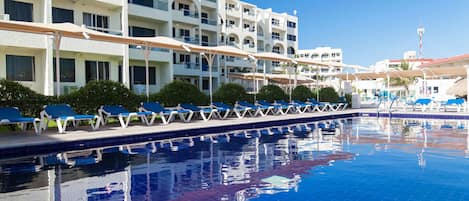 A swimming pool with blue tiles, surrounded by white buildings with balconies and blue lounge chairs.