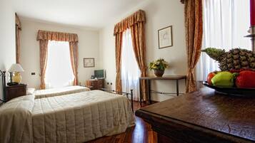 A hotel room with two beds, a wooden table with fruit, a lamp, and curtains.