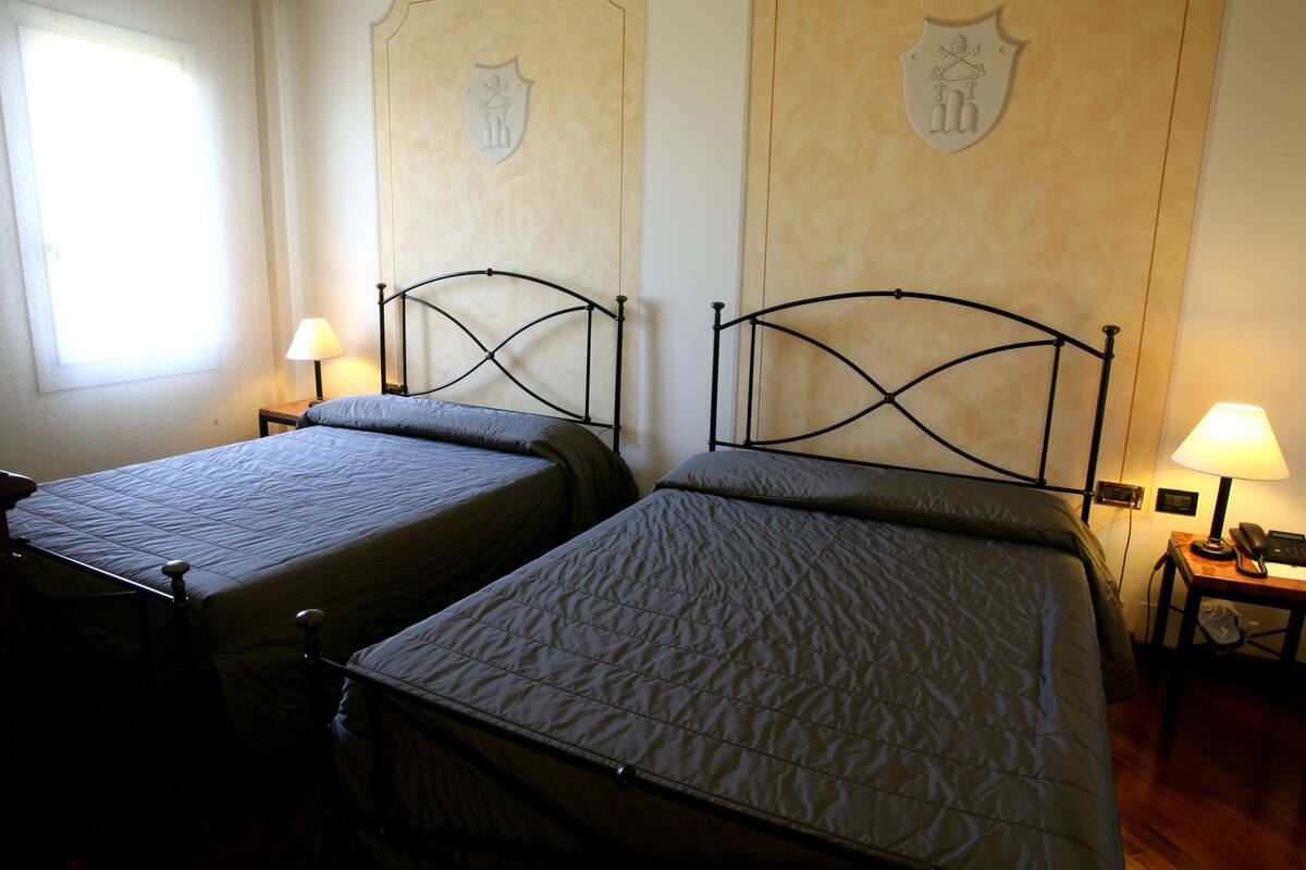 Zwei Einzelbetten mit Metallgestellen, ein Fenster mit Tageslicht und ein an der Wand befestigter Kleiderhaken.