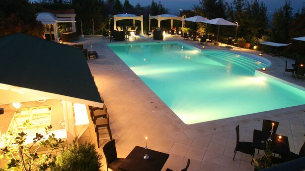 A swimming pool with surrounding lounge chairs and tables, a covered seating area, and a view of the sea. - Hotel Monte del Re (Dozza)