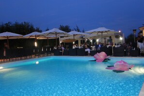 A pool area with lounge chairs and umbrellas, surrounded by a covered seating area with tables and chairs.