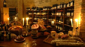 A wine cellar with shelves of wine bottles, a wooden table with wine glasses and cheese, and a stone wall. - Hotel Monte del Re (Dozza)