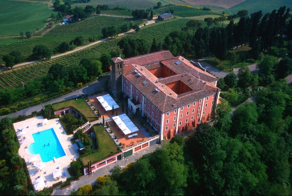 An aerial view of a large building with a swimming pool, surrounded by lush greenery and vineyards.