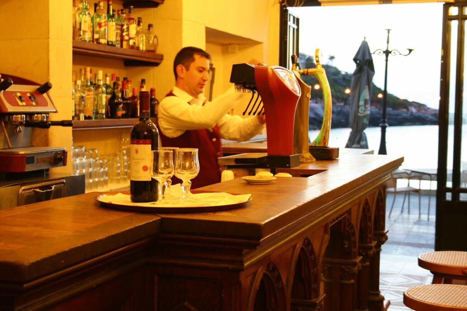 Una zona de bar con un barman preparando bebidas, una botella de vino y vasos sobre la barra.