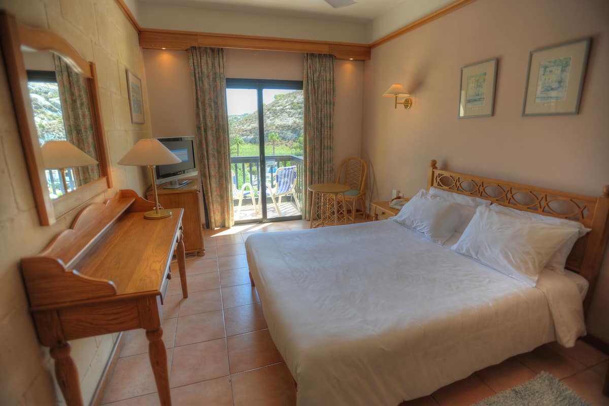 A hotel room with a bed, a desk, a chair, a TV, and a balcony with a view of greenery.