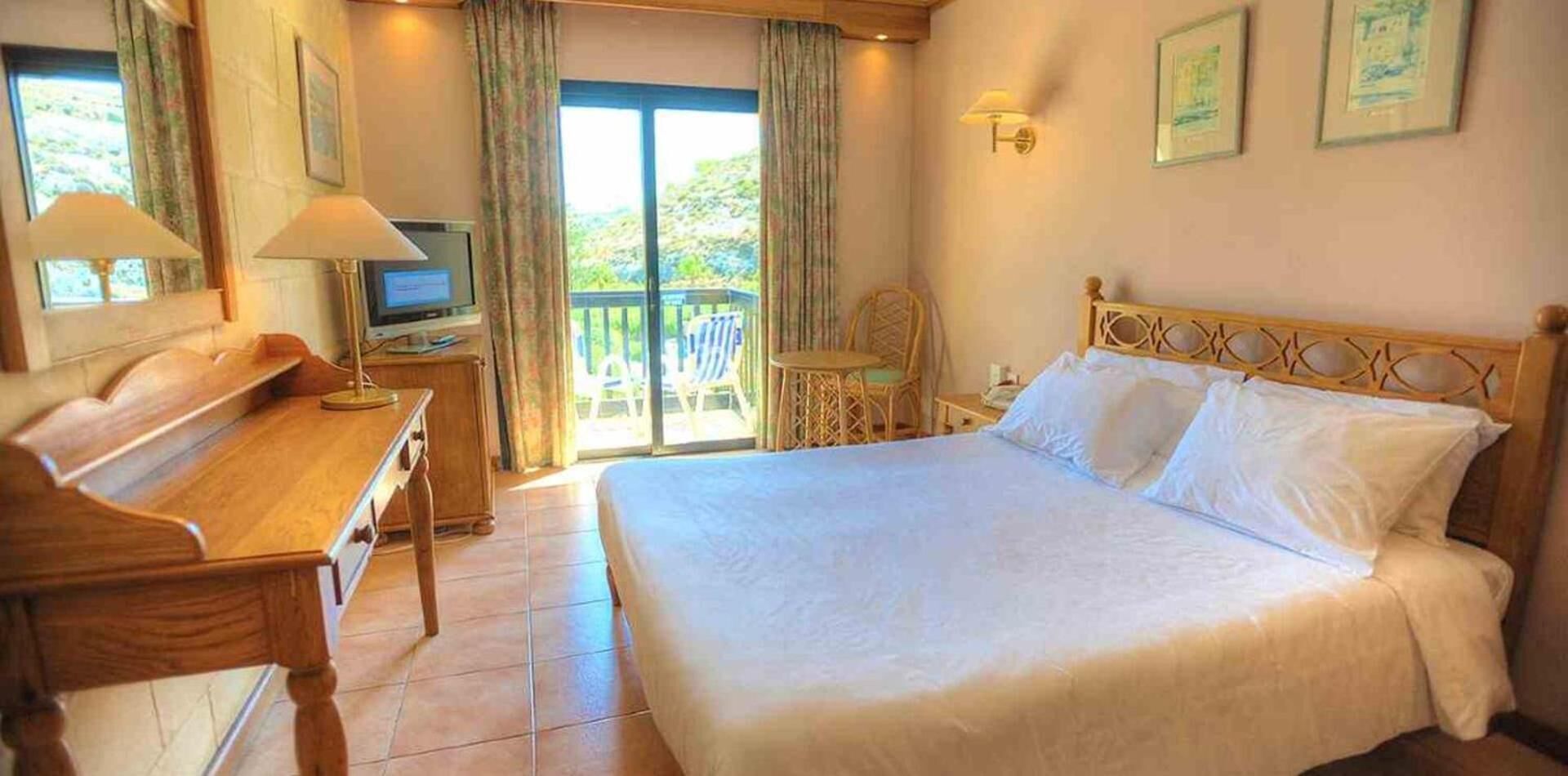 A hotel room with a bed, a desk, a television, and a balcony with chairs.