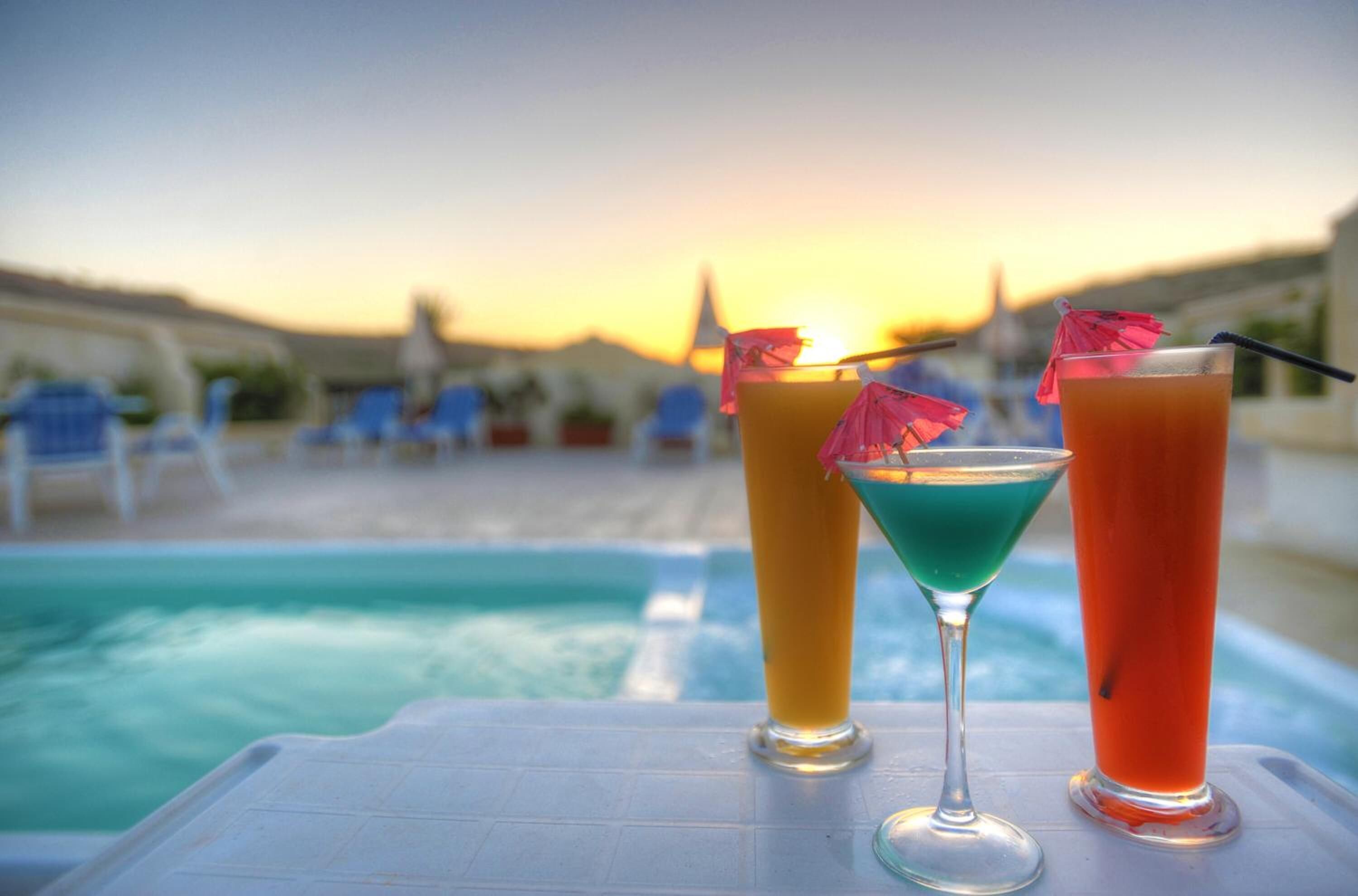 A poolside table with three drinks, a sunset in the background, and lounge chairs around the pool.