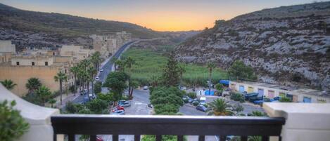 A view from a balcony overlooking a street with buildings, parked cars, and a hilly landscape.