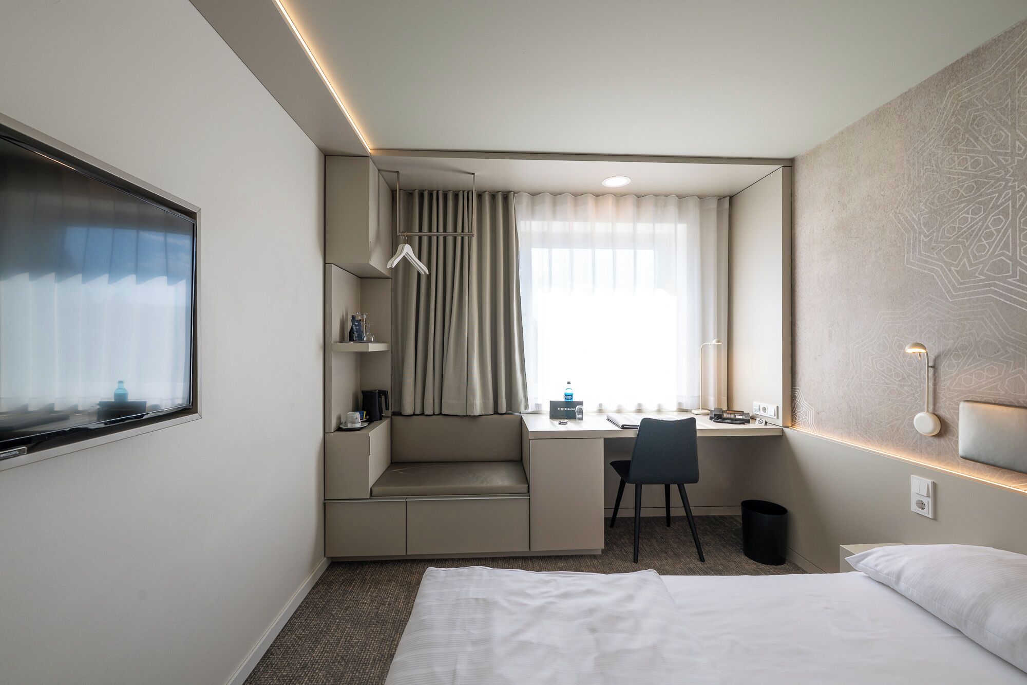 Ein Hotelzimmer mit Bett, Schreibtisch mit Stuhl, einer kleinen Sitzecke, einem Fenster mit Vorhängen und einer Wandleuchte.