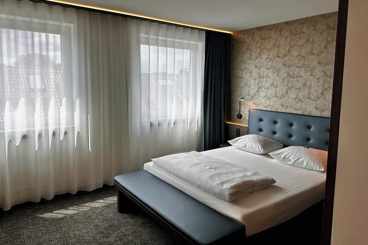 A hotel room with a large bed, a bench, a window with sheer curtains, and a patterned wall.