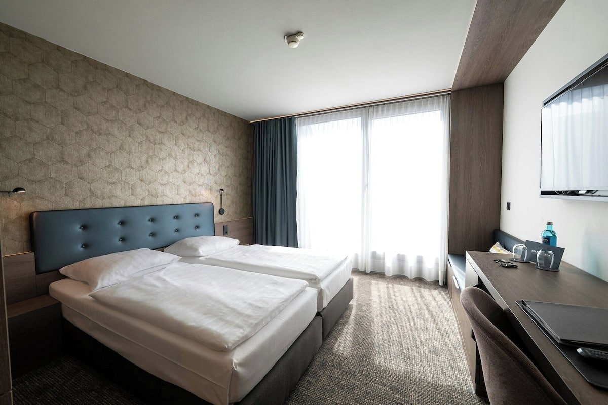 A hotel room with two beds, a desk, a chair, a TV, and a large window with curtains.