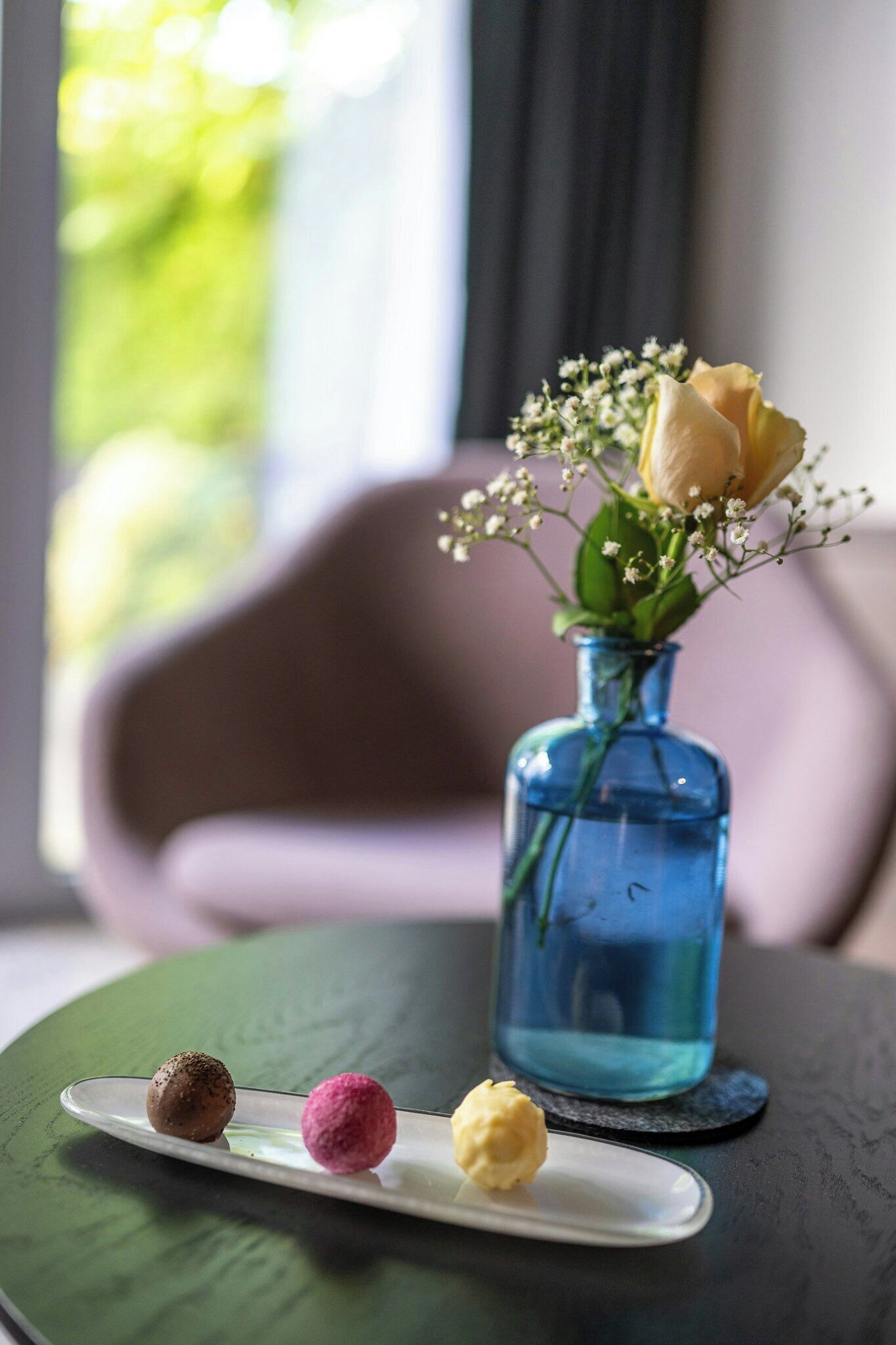 A table with a blue vase containing a yellow rose and baby's breath, and a white plate with three desserts.