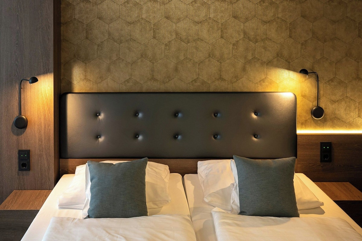 A hotel room with a large bed, a headboard with buttons, and wall-mounted lighting.