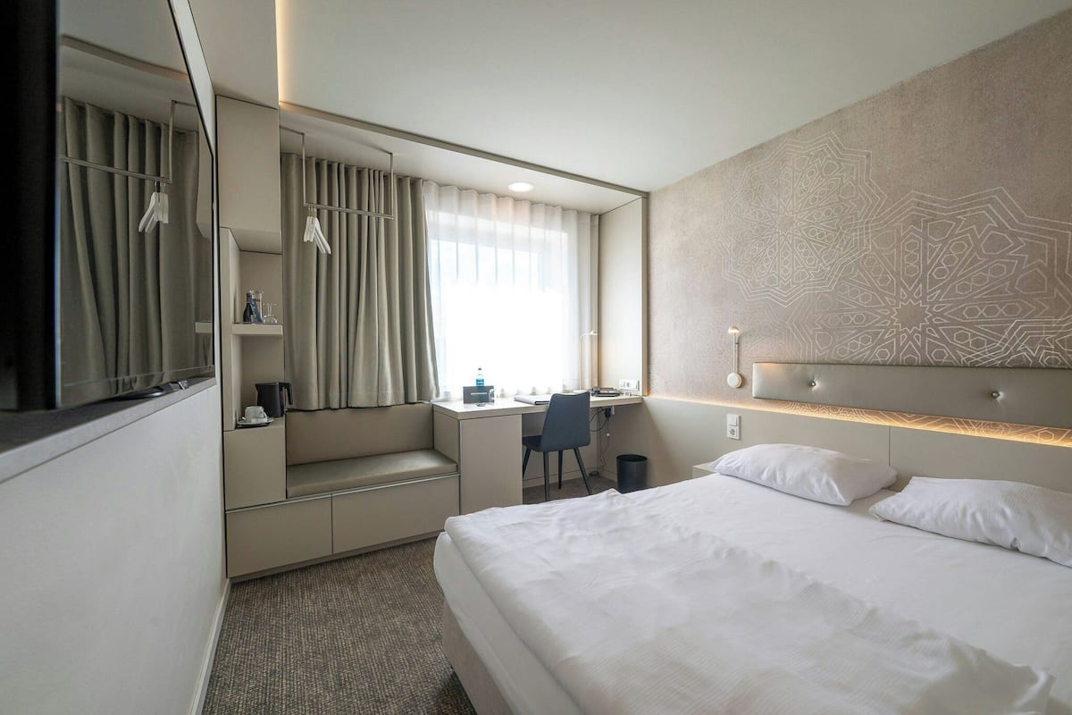 A hotel room with a bed, a desk, a chair, a mirror, and a window with curtains.