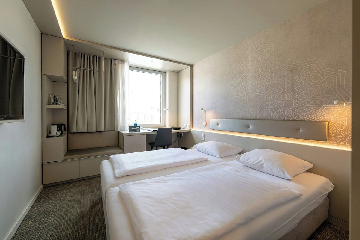 A hotel room with two beds, a desk, a chair, a window with curtains, and a decorative wall.