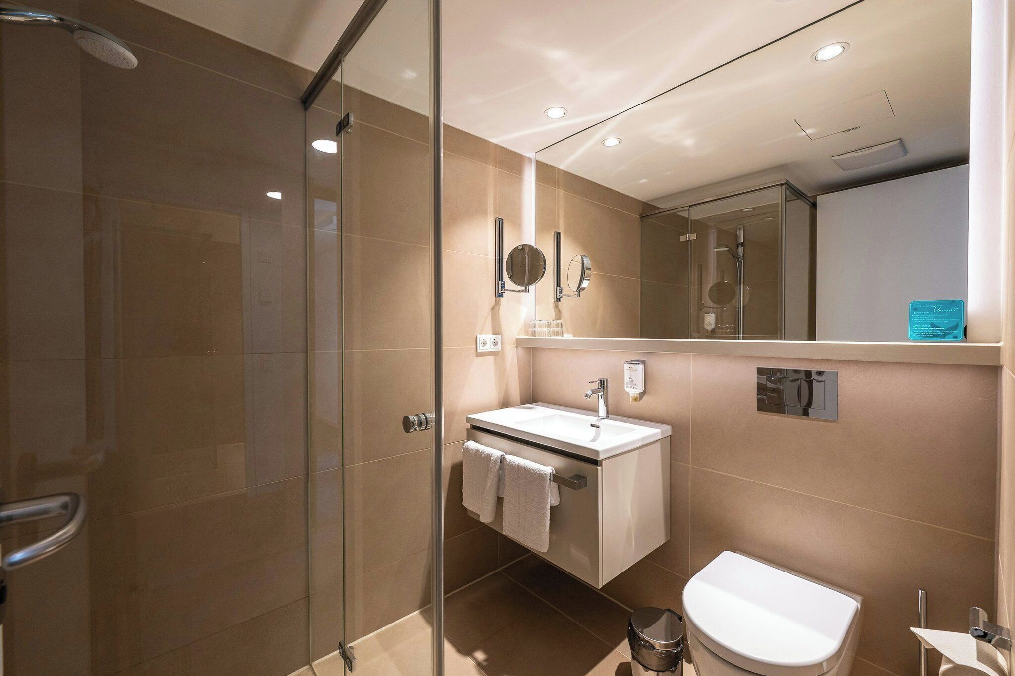 Een moderne badkamer met een douchecabine van glas, een wit wastafeltje, een spiegel en een toilet.