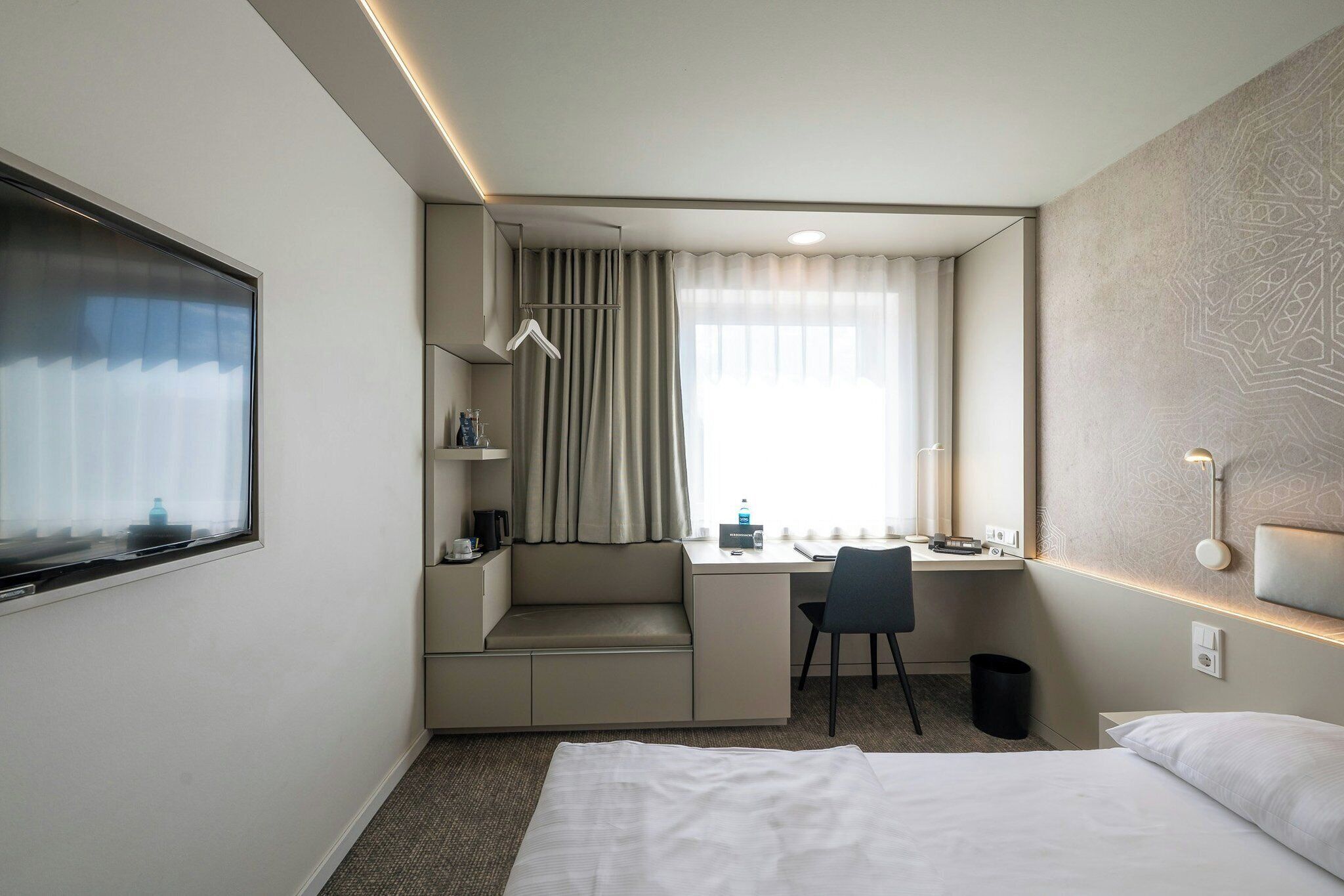 Ein Hotelzimmer mit Bett, Schreibtisch mit Stuhl, einer kleinen Sitzecke, einem Fenster mit Vorhängen und einer Wandlampe.