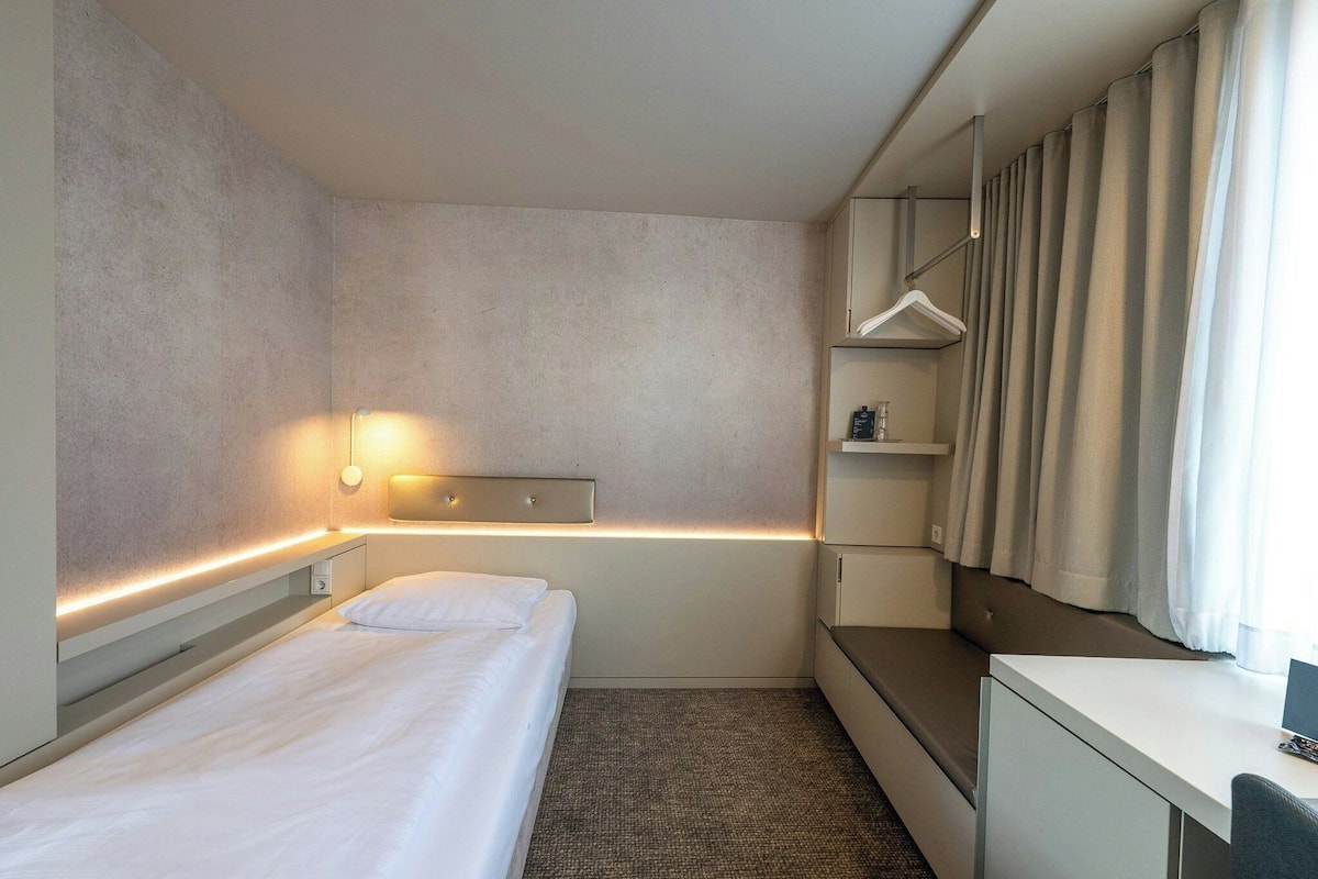 A hotel room with a bed, bedside table, wardrobe, and a desk with a chair.