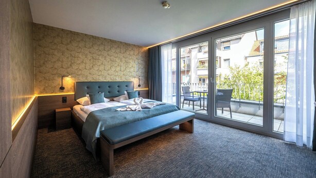 A hotel room with a large bed, a nightstand, a wall-mounted lamp, and a view of the outside balcony. - Stadthotel Münster (Münster)