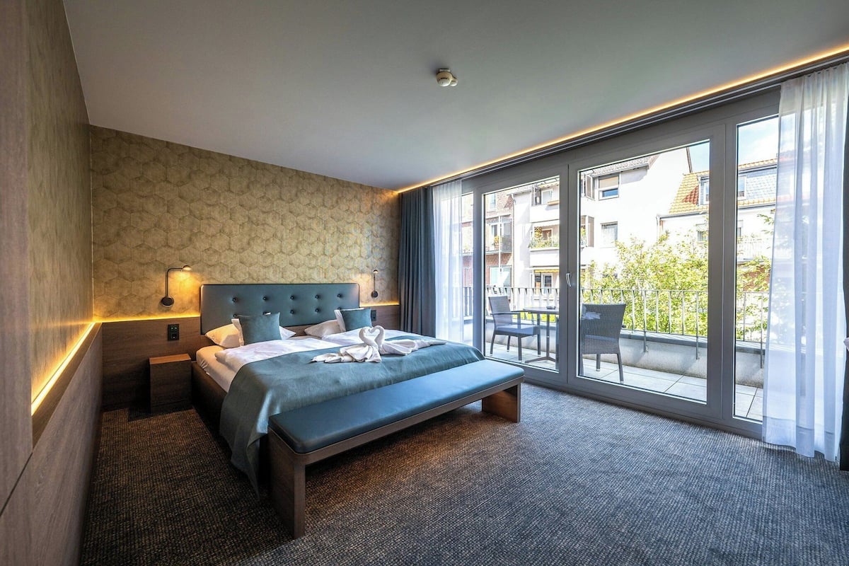 A hotel room with a large bed, a nightstand, a wall-mounted lamp, and a view of the outside balcony.