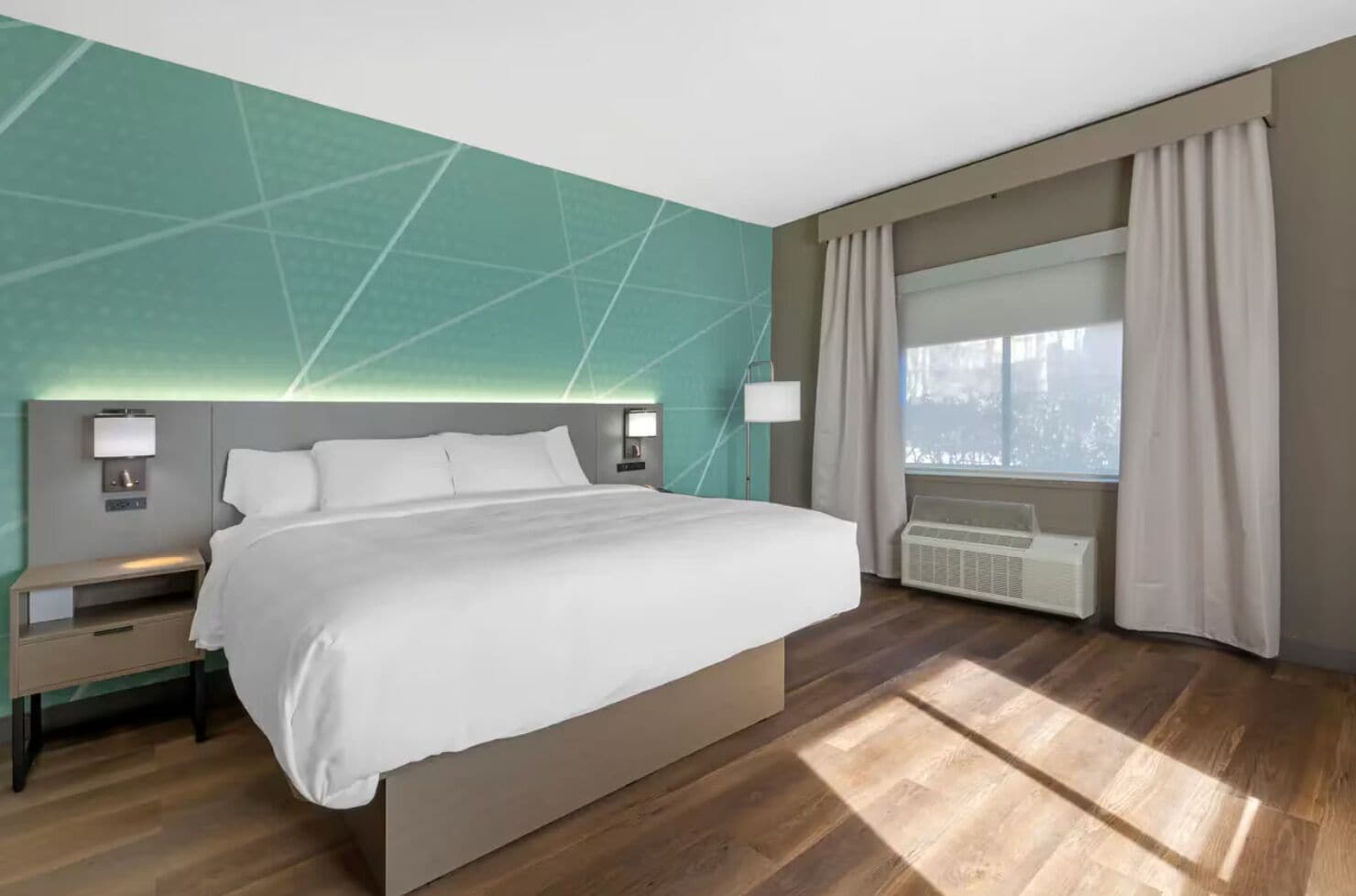 Una habitación de hotel moderna con una cama grande, mesitas de noche, una lámpara de pared, una ventana con cortinas y un equipo de aire acondicionado integrado.