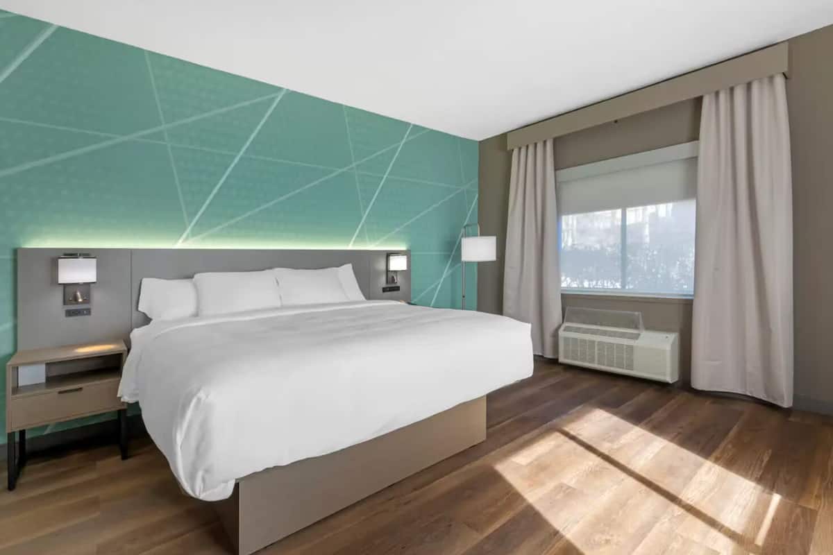 Una habitación de hotel moderna con una cama grande, mesitas de noche, una lámpara de pared, una ventana con cortinas y un equipo de aire acondicionado integrado.