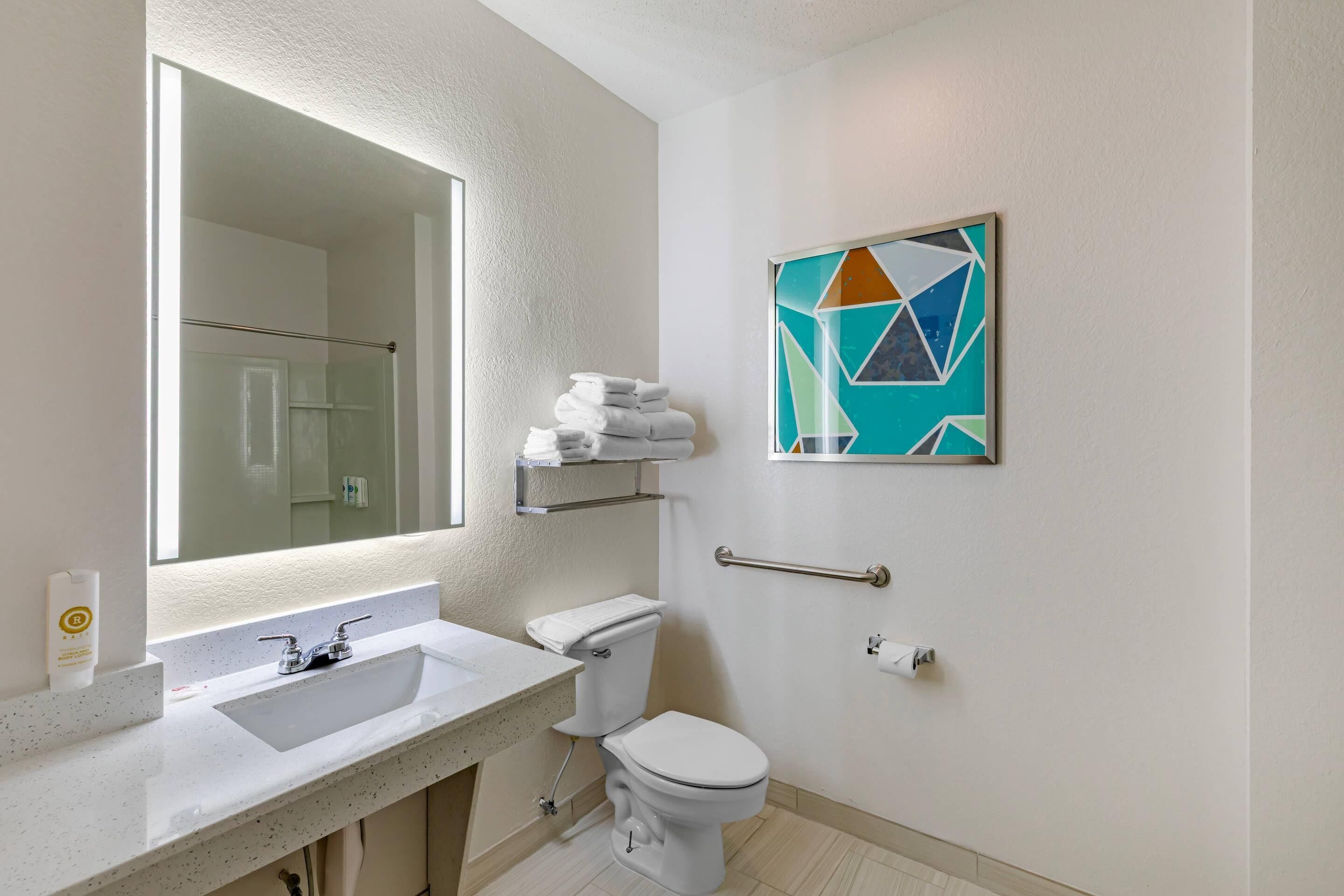 A modern bathroom with a large mirror, white sink, and a toilet. There is a towel rack above the toilet and a geometric art piece on the wall.