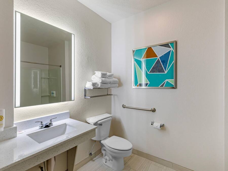A modern bathroom with a large mirror, white sink, and a toilet. There is a towel rack above the toilet and a geometric art piece on the wall.
