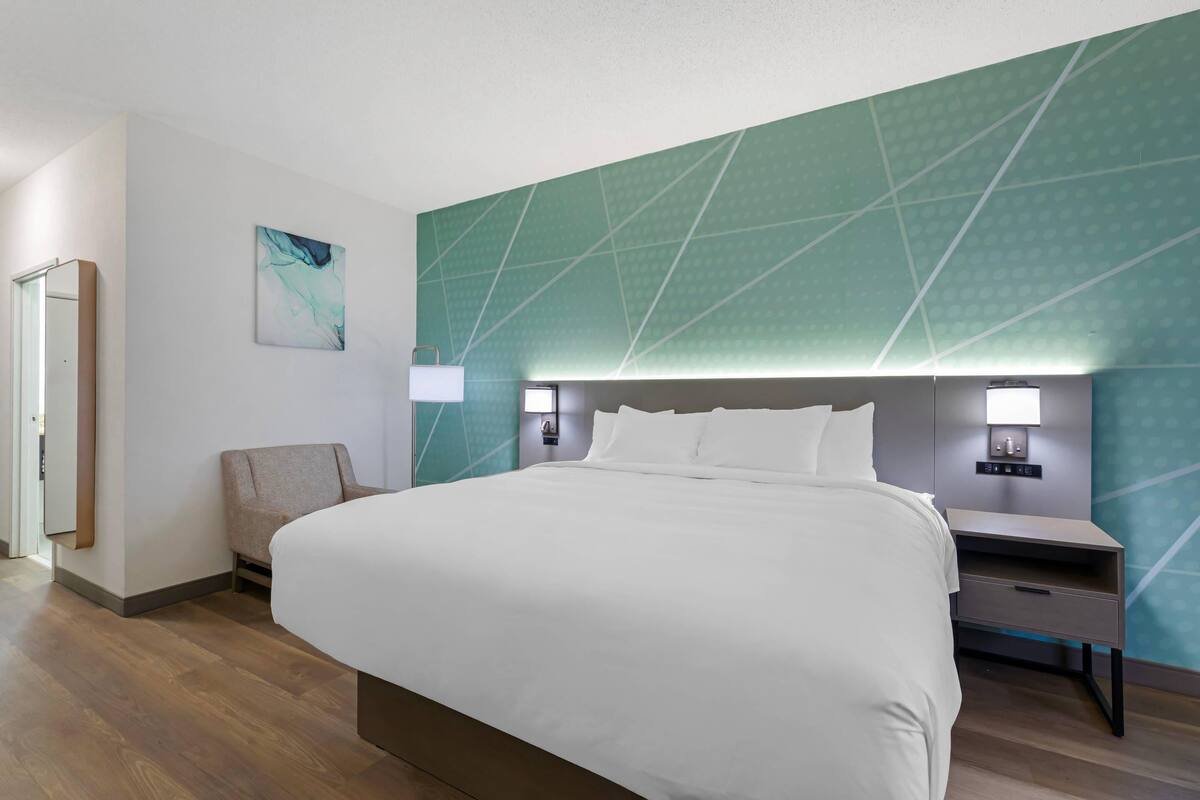 Una habitación de hotel moderna con una cama grande, una silla, una mesita de noche y una lámpara de pared.