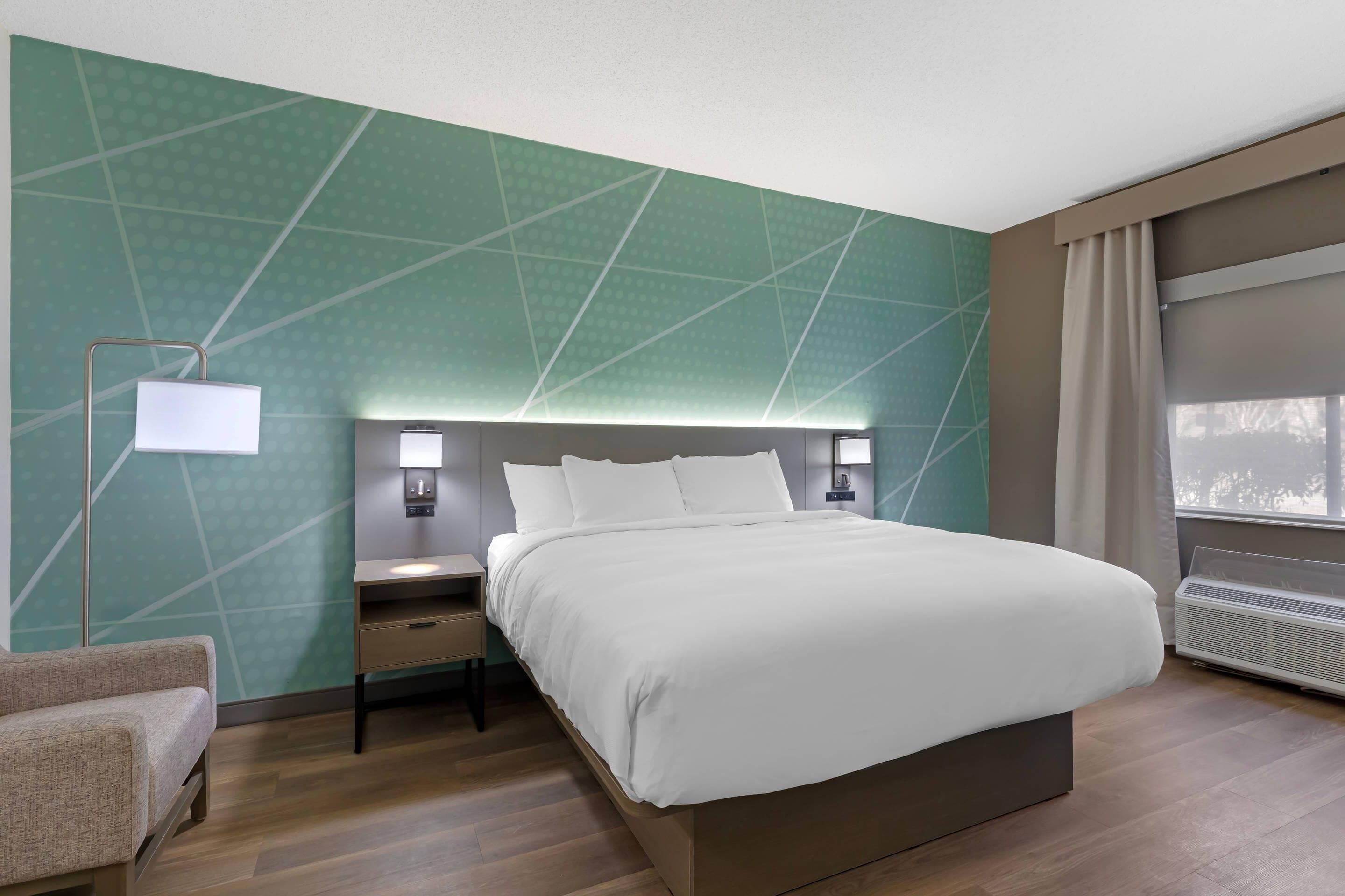 A modern hotel room with a large bed, bedside tables, a lamp, and a view of the outdoors.