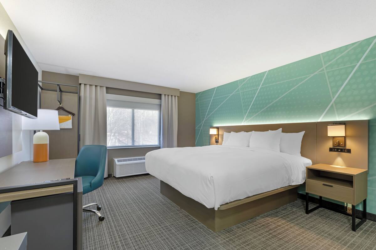 Una habitación de hotel con cama, escritorio, silla, televisor y ventana con cortinas.