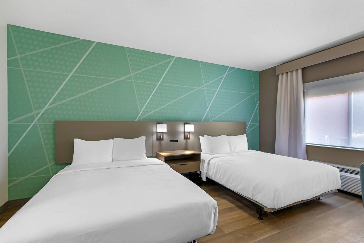 Habitación de hotel con dos camas, una mesita de noche, luces fijadas a la pared y un ventanal con cortinas.