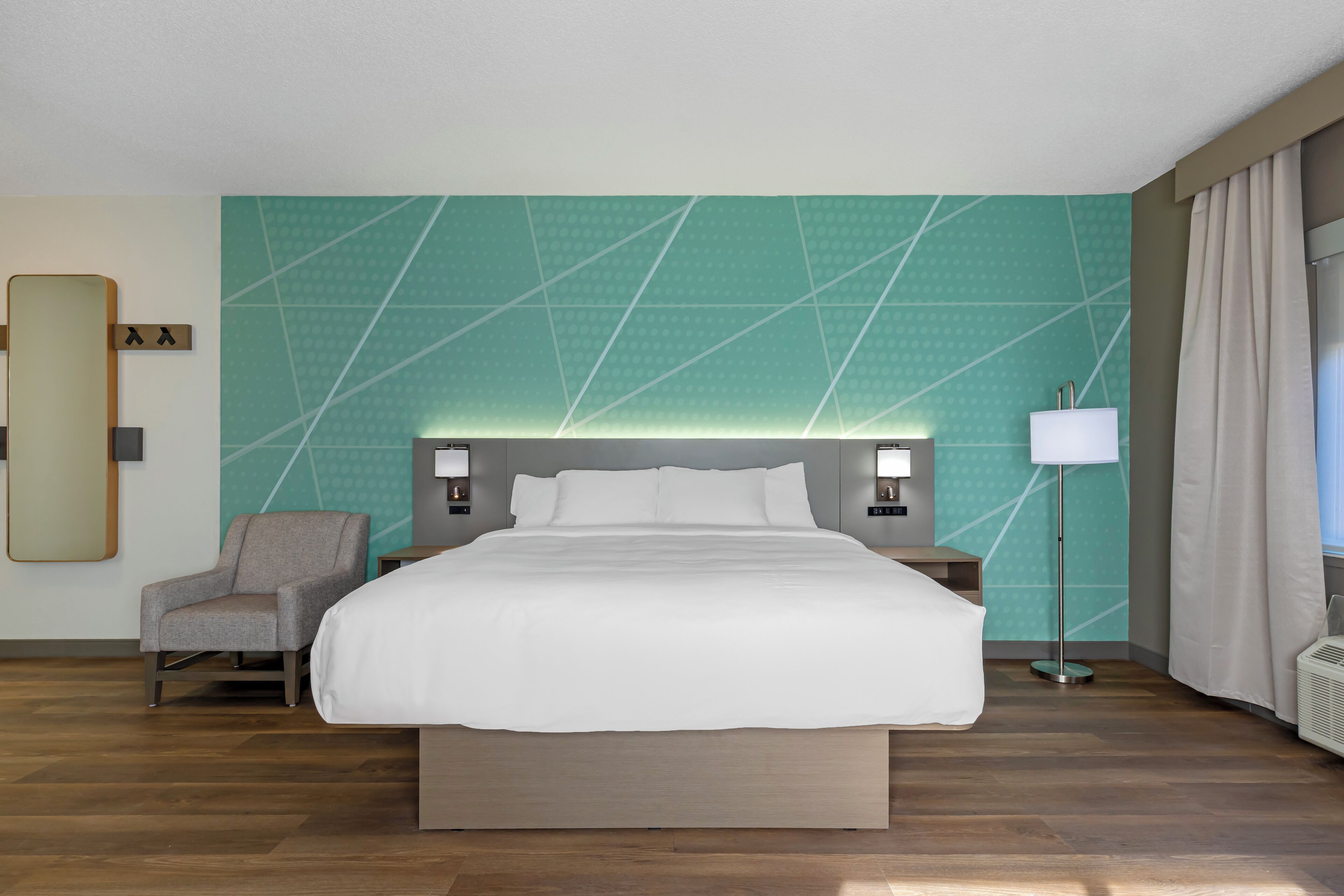 Une chambre d’hôtel avec un grand lit, une chaise, une table de chevet, une lampe et un miroir fixé au mur.