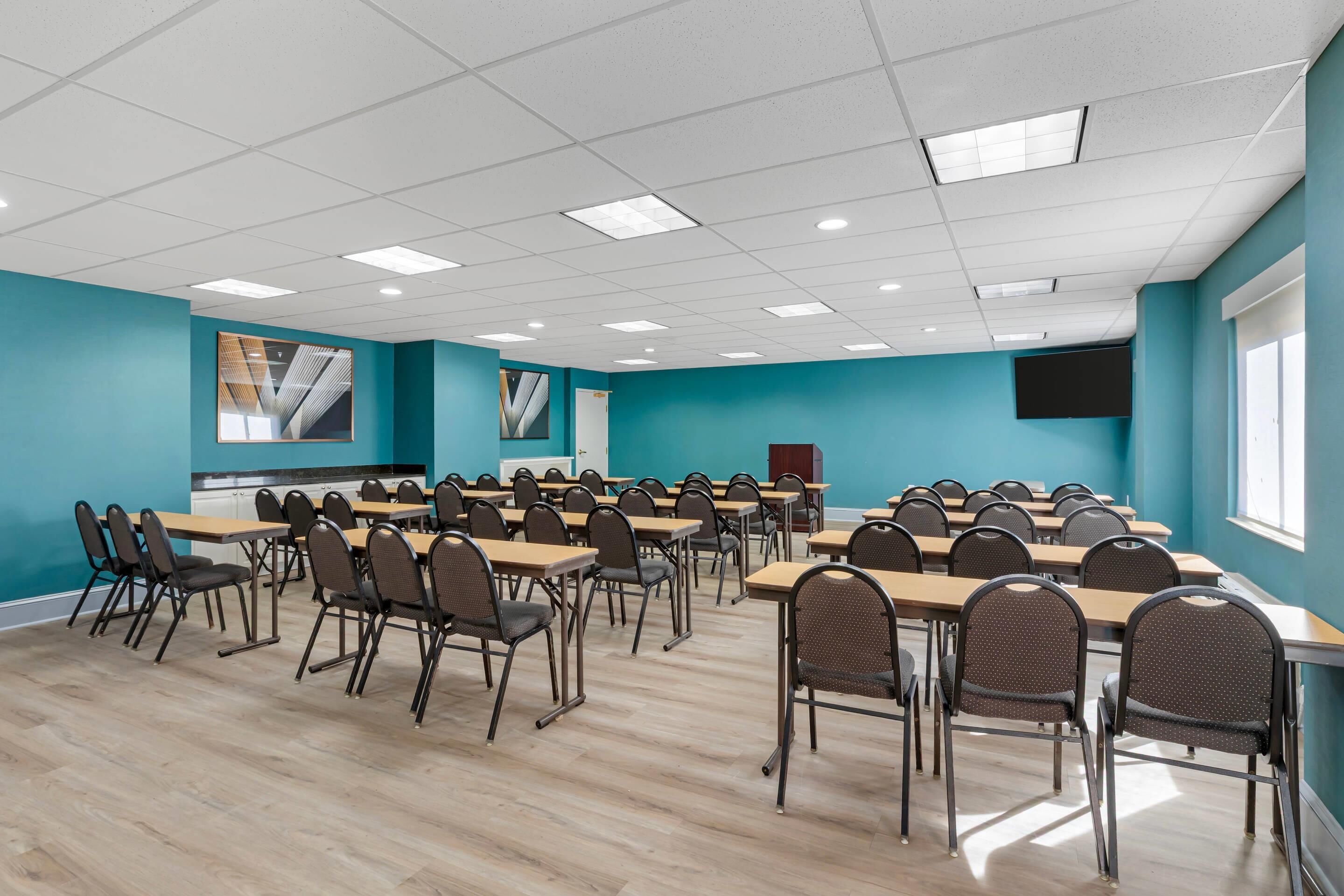 A conference room with teal walls, wooden tables, and chairs, equipped with a projector and a TV screen.