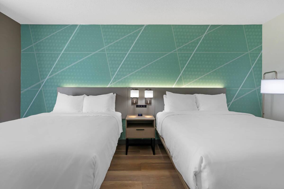 Habitación de hotel con dos camas, una mesita de noche con lámpara y una pared con diseño geométrico.