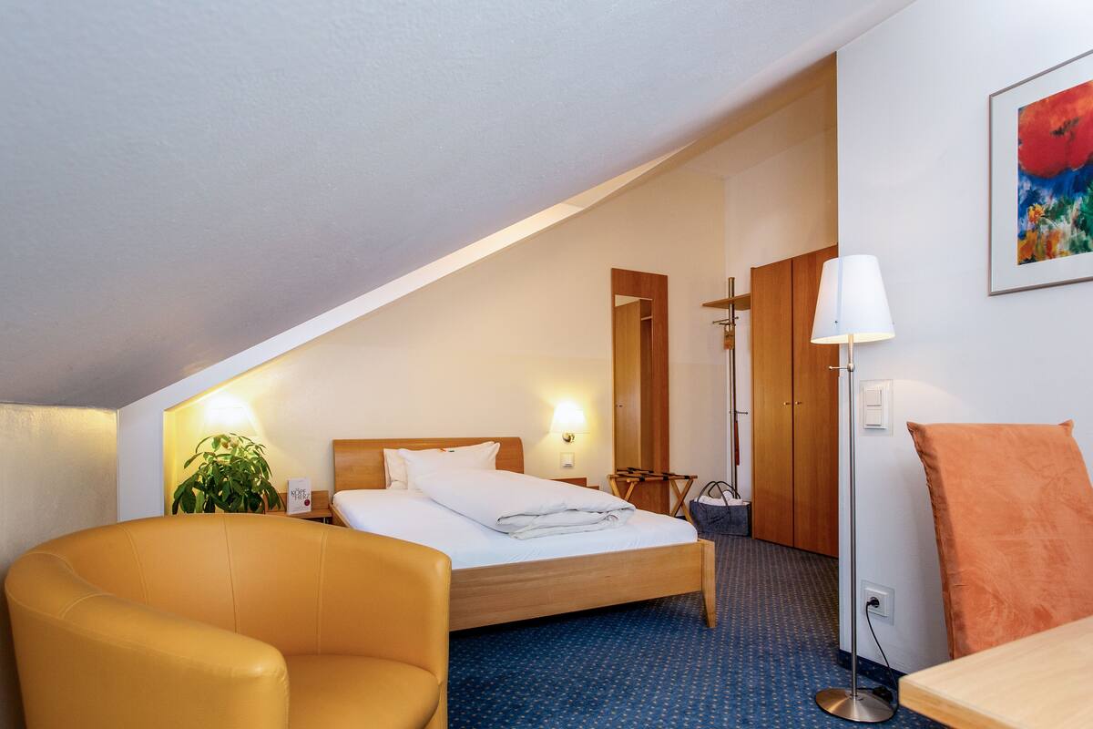 A hotel room with a bed, a chair, a desk, a lamp, a plant, and a painting on the wall.