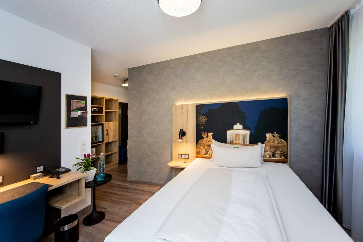 A modern hotel room with a large bed, a desk with a computer, a flat-screen TV, and a nightstand with decorative items.