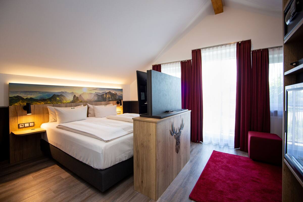 A modern hotel room with a large bed, a wooden headboard, a flat-screen TV, and a painting of mountains.
