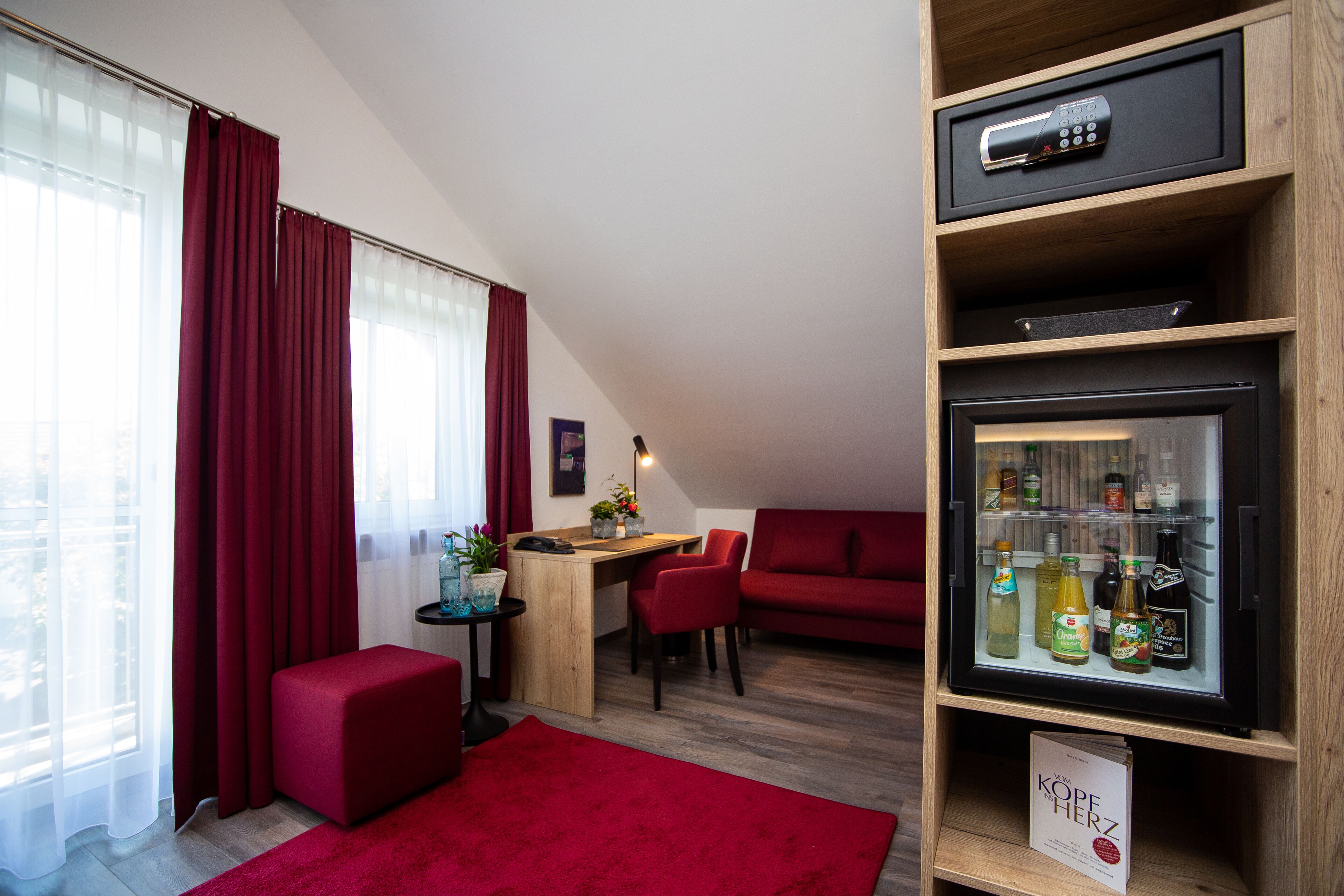 A compact living space with a wooden desk, a red sofa, a red ottoman, and a minibar with various beverages.