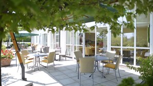 An outdoor patio with tables and chairs, large windows, and potted plants. - ARCUS HOTEL MÜNCHEN MESSE (Vaterstetten)