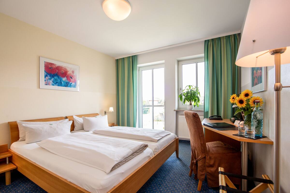 A hotel room with two beds, a desk, a chair, a lamp, a window with curtains, and a painting on the wall.