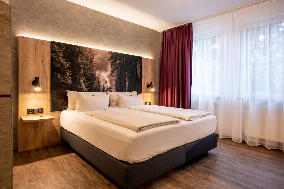A modern hotel room with a large bed, wooden headboard, and a large window with sheer curtains.