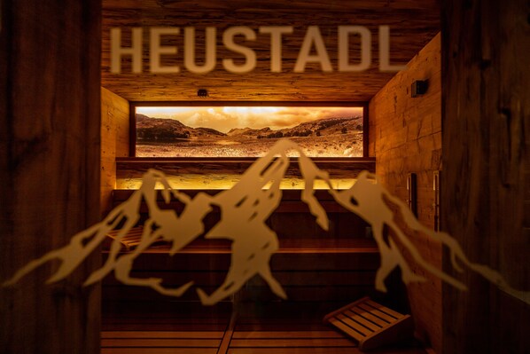 A wooden cabin interior with a large window displaying a scenic view, a bench, and a sign reading "HEUSTADL". - ARCUS HOTEL MÜNCHEN MESSE (Vaterstetten)