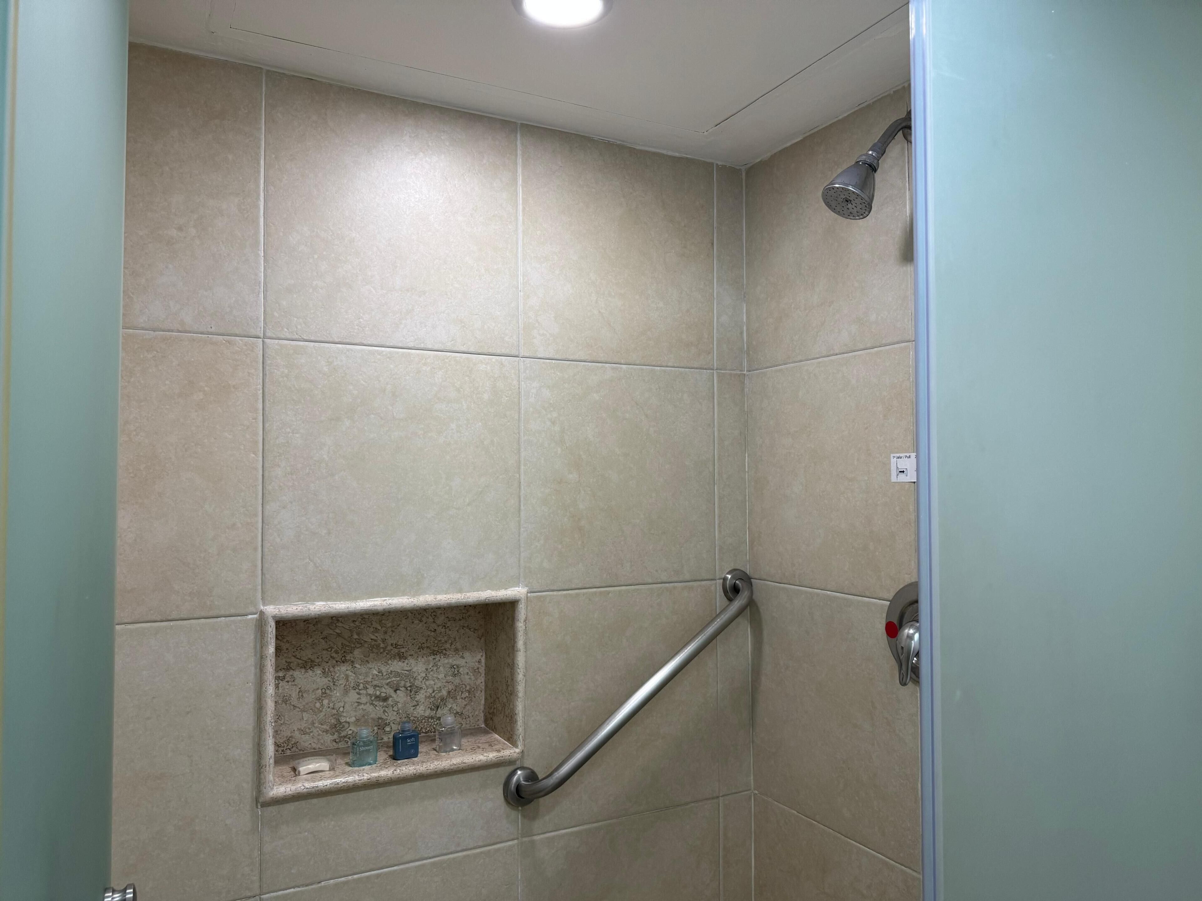 A shower area with beige tiles, a handheld showerhead, a grab bar, and a shelf with toiletries.
